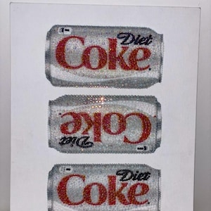 Puede incluir: Obra de arte con tres latas de Coca-Cola Light, cada una adornada con cristales brillantes. Las latas son plateadas con la inscripción "Coke" en rojo y las palabras "Diet" en negro. Fondo blanco.