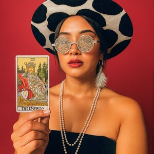 May include: A person wearing a black and white cow print hat, round glitter sunglasses, and a pearl necklace holds a tarot card that reads "THE EMPRESS". The background is a solid red.