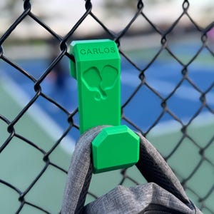 May include: A vibrant green hook, bearing the name "CARLOS", attached to a chain-link fence. The hook, featuring a pickleball paddle design, supports a grey backpack. The background reveals a pickleball court.