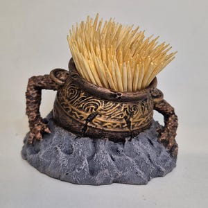 Könnte beinhalten: Ein dekorativer Zahnstocherhalter in Form eines Kessels mit Bronze-Finish und aufwendigen Details. Der Kessel steht auf einem grauen, felsigen Sockel und ist mit Holz-Zahnstochern gefüllt.