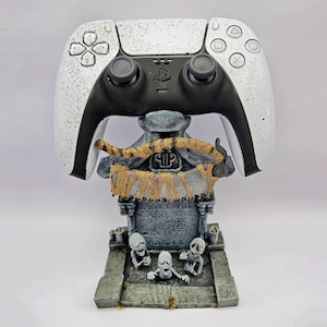 Könnte beinhalten: Ein weiß-schwarzer PlayStation 5-Controller liegt auf einem grauem Grabstein-förmigen Ständer. Der Ständer zeigt drei kleine Skelettfiguren und ein Banner mit den Buchstaben "RIP". Der Controller hat ein gesprenkeltes Design.