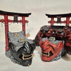 May include: Two decorative gaming controller stands featuring Japanese Oni mask designs. One is black and gray, the other red and black. Each has a red Torii gate and gold accents.