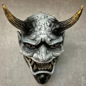 Könnte beinhalten: Eine detaillierte, handgefertigte Maske mit dämonischem Aussehen. Die Maske hat eine rissige, strukturierte Oberfläche in Grau- und Weißtönen mit schwarzen Akzenten. Sie hat große, goldene Hörner und bedrohliche Augen sowie einen weit geöffneten Mund mit sichtbaren Zähnen.
