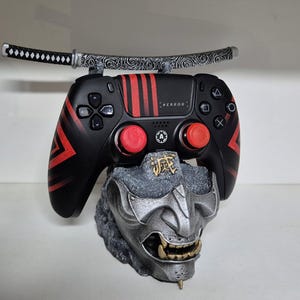Könnte beinhalten: Ein schwarzer und roter Gaming-Controller mit einem Katana-Schwert darüber. Der Controller hat rote Akzente und Knöpfe und ist auf einem grauen, detaillierten Oni-Masken-Sockel montiert. Das Wort "REDRAGON" ist auf dem Controller sichtbar.
