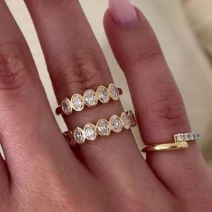 May include: A hand displays three gold rings. Two rings feature a series of bezel-set, clear gemstones. The third ring is a thin gold band with a small row of gemstones. The rings are on a hand with a light skin tone.