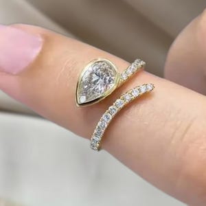 May include: A gold ring with a pear-shaped diamond center stone. The band wraps around the finger and is encrusted with small, sparkling diamonds. The ring is a piece of fine jewelry.