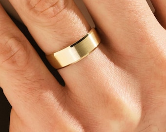 May include: A gold-colored ring with a flat, brushed surface. The ring is worn on a finger, showcasing its simple, elegant design. The ring's band is approximately 6 mm wide.