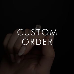 May include: A hand holding a gold ring with a large, rectangular, clear gemstone. The ring is set against a black background. The words "CUSTOM ORDER" are written in white, capital letters.
