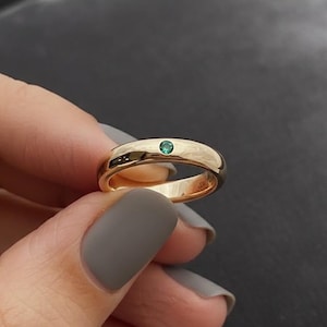 Peut inclure: Une bague en or avec une petite pierre précieuse ronde vert émeraude. La bague a une surface lisse et polie et un design de bande classique. La bague est présentée sur un fond sombre.