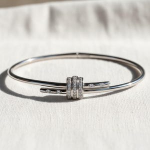 May include: A silver-toned bangle bracelet with a central design of three stacked bands encrusted with small, clear gemstones. The bracelet has a sleek, modern design with additional gemstones set along the ends.