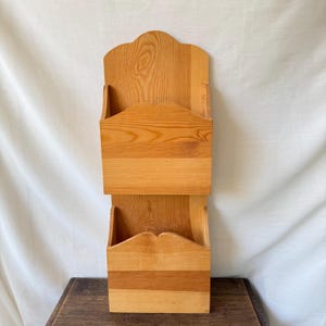 May include: A vertical wall-mounted wooden organiser featuring two stacked open compartments. The unit is crafted from light-toned wood with a visible natural grain pattern, featuring a scalloped top edge and curved cutouts on the front panels.
