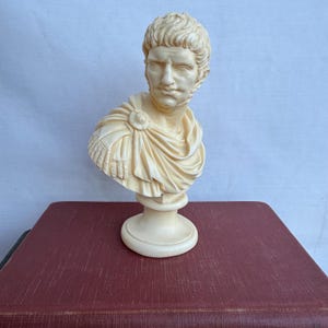 May include: Ivory-colored bust of a Roman emperor, featuring detailed facial features and a draped toga. The sculpture is mounted on a pedestal base, showcasing classical art and historical decor.