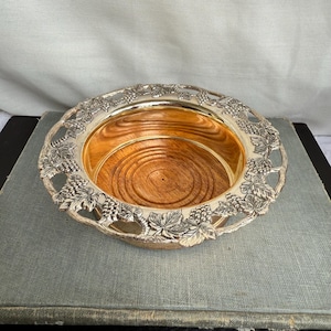 May include: Silver-toned decorative bowl with a wooden interior. The rim features a detailed grape and leaf design. The bowl has a circular shape and is likely used for decorative purposes or serving.