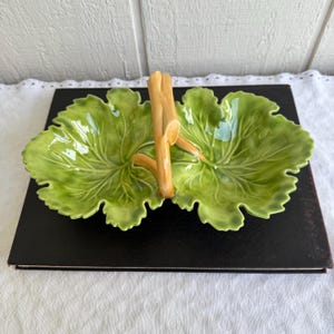 May include: A green ceramic serving dish shaped like two overlapping leaves, with detailed veining. A light brown handle is attached in the center. The dish is on a black surface, with a white background.