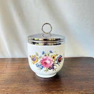May include: A white ceramic canister featuring a colorful floral pattern with a large central pink rose, surrounded by smaller blue, yellow, and purple flowers. The container has a polished silver-tone metal lid with a circular ring handle.