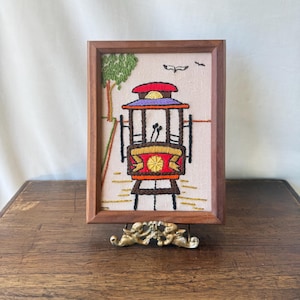 May include: A framed embroidery art piece featuring a red and brown cable car on a beige fabric background. The scene includes a green tree on the left, a small bird in the sky, and a yellow circular emblem on the front of the cable car.