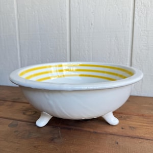 Può includere: Ciotola in ceramica bianca con tre piedini. Il bordo interno presenta strisce gialle orizzontali. La ciotola è appoggiata su una superficie di legno contro uno sfondo di legno bianco. La ciotola ha un'estetica classica e vintage.