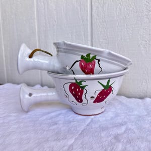 May include: Two white ceramic colanders with handles, stacked. The colanders are decorated with hand-painted red strawberries with green leaves. The colanders have small drainage holes and a grey stripe around the rim.