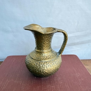 May include: A vintage brass jug with a textured surface and a handle. The jug has a rounded base and a spout. The brass has a warm, golden colour. The jug is likely used for decorative purposes.