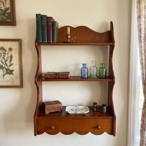 May include: A three-tiered wooden wall shelf with a scalloped top and bottom edge, featuring a single drawer at the base. The shelves hold four books, a brass candle holder, three glass bottles, a leather-bound book, a wooden box, and a dish.