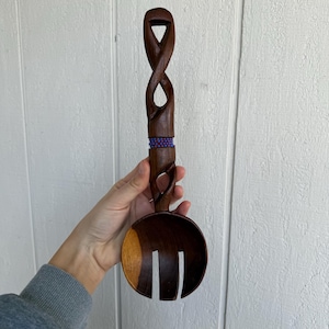 Puede incluir: Una cuchara de ensalada de madera marrón oscuro con un mango tallado con un diseño de símbolo de infinito. El mango está adornado con una banda de pequeñas cuentas moradas y azules. La cuchara tiene un cuenco ranurado.