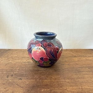 Puede incluir: Un pequeño jarrón de cerámica redondeado con un borde azul oscuro y un colorido patrón en relieve de granadas rojas, uvas moradas y hojas verdes estilizadas. La superficie tiene un acabado brillante con tonos azul profundo, rojo y morado.