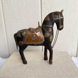 Pode incluir: Estatueta decorativa de cavalo de madeira com sela e freio. O cavalo é marrom escuro com detalhes metálicos dourados e acobreados. A sela é cor de cobre com um desenho floral. Uma corrente de metal serve como freio.