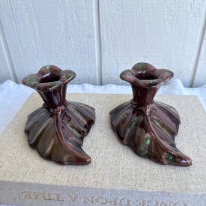 May include: Two ceramic candle holders in a leaf design. The candle holders are a deep burgundy color with green accents. The base of each candle holder is shaped like a leaf. The candle holders are sitting on a book.