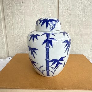 Puede incluir: Un tarro de jengibre de cerámica blanca con un diseño de bambú azul. El tarro tiene un cuerpo redondeado y una tapa a juego. Los tallos y hojas de bambú están pintados con un patrón estilizado y detallado. El tarro está sobre una superficie marrón.