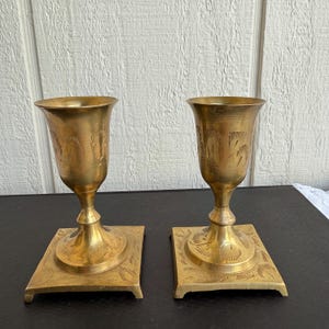 May include: Two vintage brass goblets with etched floral designs. Each goblet has a flared rim, a slender stem, and a square base. The brass has a warm, golden hue, and the goblets are displayed on a dark surface.
