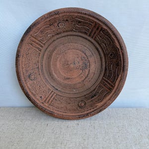 Puede incluir: Un plato redondo de color terracota con un diseño en relieve. El plato presenta círculos concéntricos y patrones decorativos de líneas onduladas y rectángulos. El borde del plato tiene un borde ligeramente elevado.