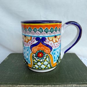 May include: A hand-painted ceramic mug with a vibrant, colorful design. The mug features a white handle with blue trim and is adorned with floral and geometric patterns in orange, blue, green, and yellow. The mug is approximately 4 inches tall.