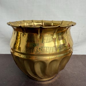 May include: A vintage brass planter with a scalloped rim and a rounded body. The planter has a shiny, reflective surface and a decorative, ridged pattern around the base. The overall shape is reminiscent of a classic flower pot.