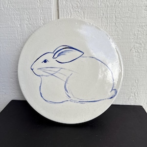 Könnte beinhalten: Ein weißer Keramikteller mit einer blauen Skizze eines Kaninchens. Das Kaninchen ist im Profil dargestellt, mit langen Ohren und einem abgerundeten Körper. Der Teller ist rund und hat einen leicht erhöhten Rand. Das Design ist schlicht und elegant.