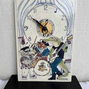 May include: Rectangular ceramic clock featuring a band of animal musicians. The clock face has a musical note and a yellow sun. An elephant plays the saxophone, a tiger drums, and an alligator plays guitar.