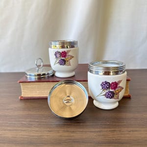 May include: Two white porcelain egg coddlers with silver-tone metal rims and lids, each decorated with a botanical illustration of purple and red blackberries with green leaves. One lid is placed upside down to show the interior metal finish.