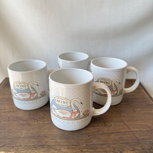 May include: A set of four white ceramic mugs featuring a repeating pattern of small blue dots and a central illustration of a white swan with a pink ribbon. Each mug displays the word HOME in a decorative oval frame above the swan illustration.