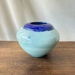 May include: A round ceramic vase featuring a pale mint green body with a fine crackle glaze finish. The wide, circular opening at the top is finished with a deep, vibrant cobalt blue glaze that contrasts with the lighter base of the vessel.