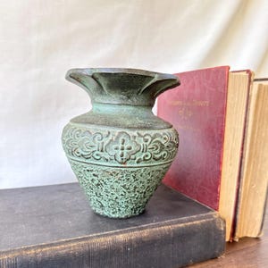 Könnte beinhalten: Eine kleine, verwitterte grüne Metallvase mit einem ausgestellten, gewellten Rand und einer texturierten, kupfergrünen Oberfläche. Der Körper zeigt ein zentrales Band mit geprägten Blumen- und Rankenmustern sowie eine raue, körnige Basis.