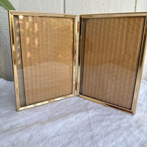 May include: A gold-colored, hinged picture frame, open to display two photos. The frame has a simple, elegant design with a thin border and a textured edge. The background is a light brown color.