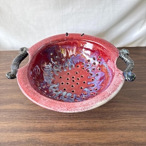 Può includere: Uno scolapasta in ceramica con una vivace finitura smaltata rossa e viola e una consistenza maculata. La ciotola presenta numerosi fori di drenaggio sulla base e sui lati, completati da due manici scuri, strutturati e ricurvi.