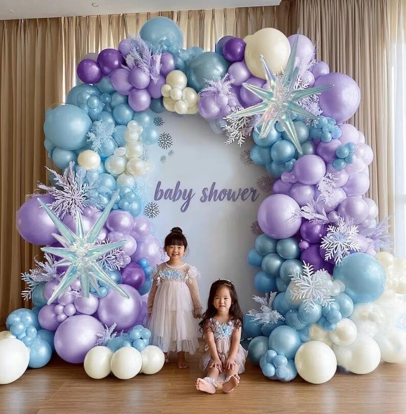 Puede incluir: Un tel&oacute;n de fondo de baby shower con un arco de globos en tonos pastel azul, morado y blanco. El arco enmarca las palabras "baby shower" y est&aacute; decorado con detalles de copos de nieve y adornos en forma de estrella. Dos ni&ntilde;as con vestidos est&aacute;n frente al tel&oacute;n de fondo.