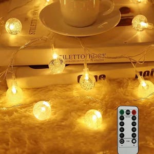 May include: A string of warm white LED globe lights with textured, bubble-like surfaces. The lights are arranged around a white ceramic cup and saucer resting on two stacked books. A rectangular remote control with numbered mode buttons is included.