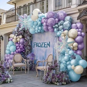 Puede incluir: Decoraci&oacute;n de fiesta tem&aacute;tica de Frozen con un arco de globos en tonos azules, morados y blancos. El arco enmarca un tel&oacute;n de fondo con la palabra "FROZEN" y est&aacute; decorado con adornos en forma de estrella y arreglos florales. Dos sillas y una mesa peque&ntilde;a est&aacute;n dispuestas delante.