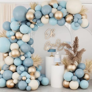 May include: A baby shower display with a balloon arch in blue, gold, and cream. The arch surrounds a backdrop with the words "Oh Baby" and two small cakes on pedestals. Dried grasses and more balloons create a celebratory atmosphere.