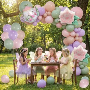 K&ouml;nnte beinhalten: Eine bezaubernde Tee-Party im Freien. Vier junge M&auml;dchen in Feenkost&uuml;men sitzen an einem kleinen Holztisch. Ein Ballonbogen in Pastellfarben und ein Teekannen-Ballon schm&uuml;cken die Szene. Ballons in verschiedenen Farben sind verstreut.