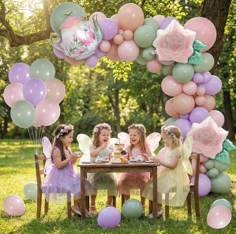 K&ouml;nnte beinhalten: Eine bezaubernde Tee-Party im Freien. Vier junge M&auml;dchen in Feenkost&uuml;men sitzen an einem kleinen Holztisch. Ein Ballonbogen in Pastellfarben und ein Teekannen-Ballon schm&uuml;cken die Szene. Ballons in verschiedenen Farben sind verstreut.