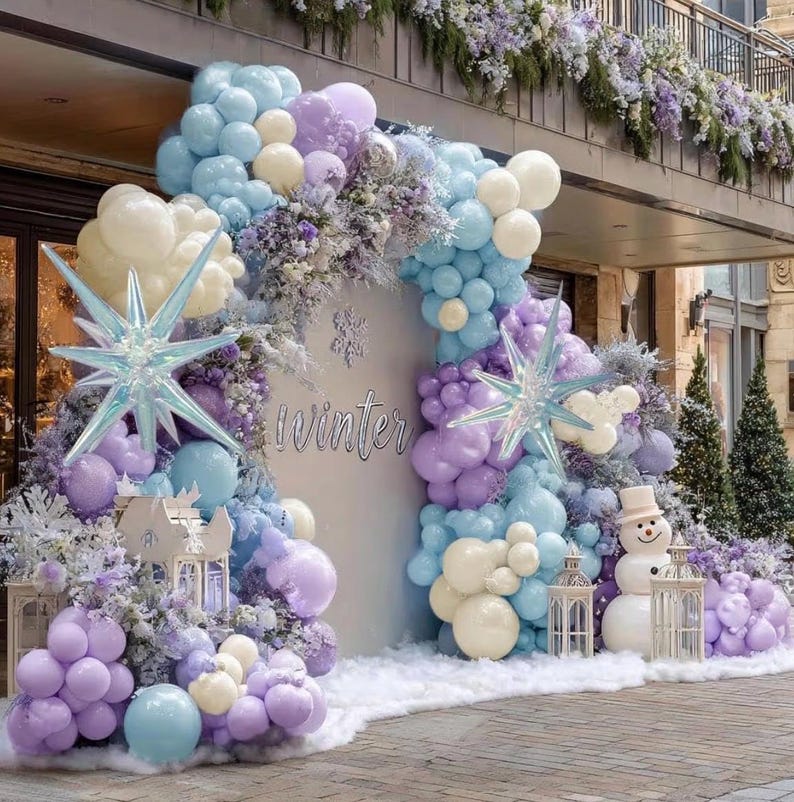 Puede incluir: Una exhibici&oacute;n con tem&aacute;tica invernal con un tel&oacute;n de fondo con la palabra "Winter". La exhibici&oacute;n incluye un arco de globos en tonos azules, morados y blancos, decoraciones en forma de estrella y un mu&ntilde;eco de nieve.