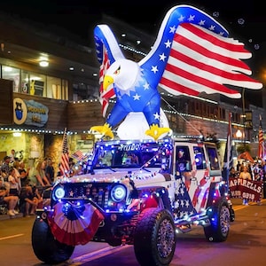 May include: A white Jeep Wrangler decorated with American flag patterns and colorful string lights, featuring a large inflatable bald eagle with spread wings mounted on the roof, displaying the word Fathom above the windshield at night.