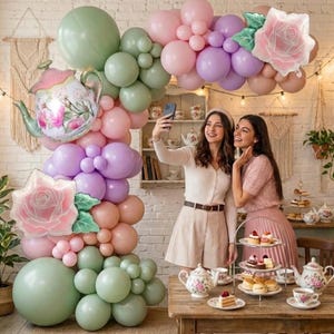 K&ouml;nnte beinhalten: Eine verspielte Party-Szene mit einem Ballonbogen in Pastellfarben, darunter Rosa, Lila und Gr&uuml;n. Ein Teekannen-Ballon und ein rosenf&ouml;rmiger Ballon erg&auml;nzen die Dekoration. Zwei Personen machen ein Selfie bei einer Teeparty.
